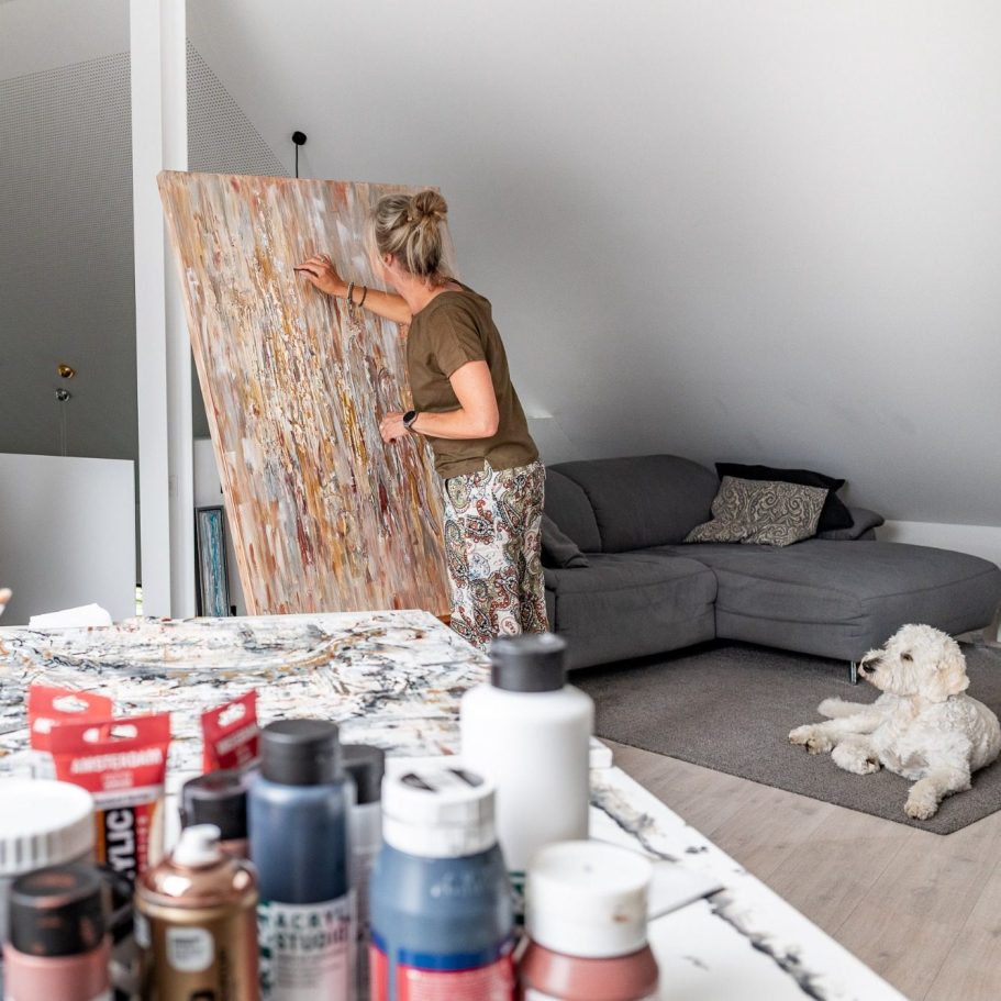 Eine Frau malt an einer Leinwand, während ein Hund vor dem Sofa sitzt.